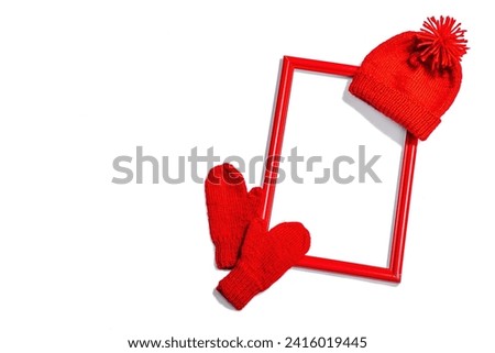 Creative handmade winter flat lay. Red wooden frame, knitted mittens and hat, cozy mood concept. Hard light, dark shadow, white background, top view
