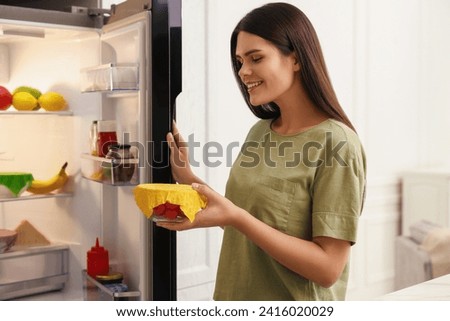 Woman holding bowl of fresh tomatoes covered with beeswax food wrap near refrigerator in kitchen