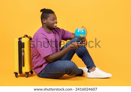 Vacation Abroad. Happy African American Traveler Man Holds World Globe Sitting Near Yellow Suitcase, Choosing Travel Destination Over Orange Studio Background. Globetrotter Concept Royalty-Free Stock Photo #2416024607