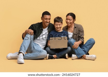 Beautiful family of three father, mother and school-aged son sitting on floor over beige studio background, watching movie or cartoon together, websurfing, using laptop computer