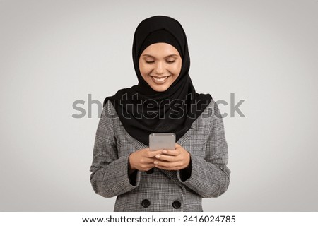 Smiling woman wearing hijab and business suit engrossed in sending message on her smartphone, embodying modern communication in style