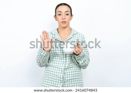Serious Beautiful young woman wearing green plaid pyjama and holding a cup pulls palms towards camera, makes stop gesture, asks to control your emotions and not be nervous