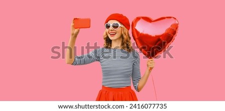 Summer portrait of happy smiling young woman taking selfie with mobile phone holding red heart shaped balloon wearing french beret, sunglasses on pink studio background