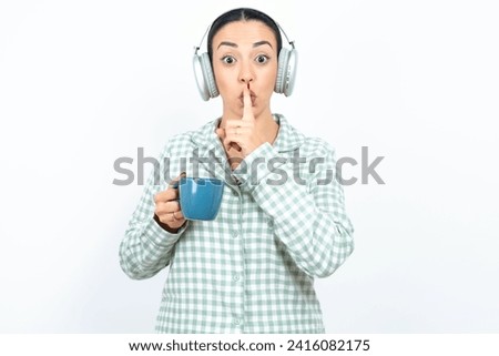 Beautiful young woman wearing green plaid pyjama and holding a cup making hush gesture with finger on her lips wearing  wireless headphones. Be quiet. Royalty-Free Stock Photo #2416082175