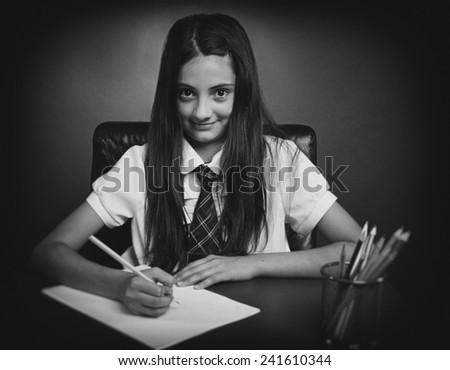 Smiling schoolgirl sits at a school desk writes homework smiling at camera. Black & white picture.