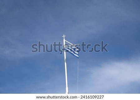 The Blue and White Striped Greek National Flag, flys at the top of a White Flagpole that has a White Cross on top of it. The background is just Blue Sky and thin White Clouds on a sunny day.