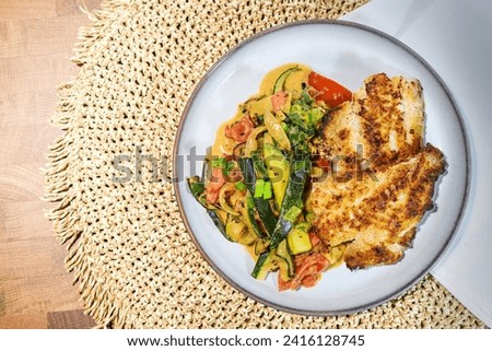 Two fried redfish fillets on vegetables such as zucchini, tomatoes and leek, served on a gray plate and a woven natural fiber placemat, healthy recipe, low carb diet, top view from above, copy space  Royalty-Free Stock Photo #2416128745