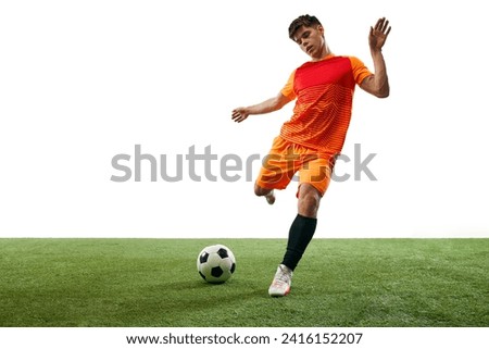 Young man, football player in orange uniform playing, training, hitting ball isolated over white background with grass flooring. Concept of sport, game, competition, championship, active lifestyle