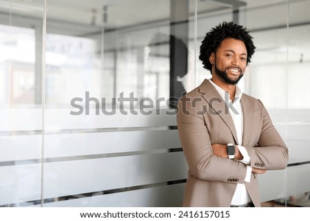 An African-American businessman stands confidently in a modern office environment, arms crossed, exuding a sense of leadership and the calm assurance of a seasoned professional Royalty-Free Stock Photo #2416157015