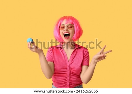 Beautiful young woman in pink wig with blue sweet macaroon showing victory gesture on yellow background Royalty-Free Stock Photo #2416262855
