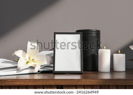 Blank funeral frame, burning candles, mortuary urn and lily flower on wooden cabinet against grey wall