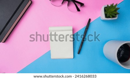 workspace desk with notebooks, pens, glasses, plants, and coffee on blue and pink background