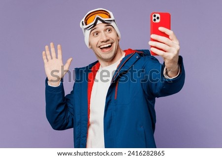 Skier man wear warm blue windbreaker jacket ski goggles mask hat do selfie shot on mobile cell phone waving hand spend extreme weekend winter season in mountains isolated on plain purple background