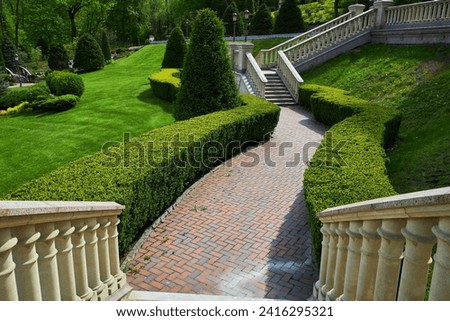 Trimmed green bushes form a labyrinth along tiled pathways in the park, creating a serene and maze-like atmosphere. A perfect summer day unfolds in this beautifully landscaped outdoor space. Royalty-Free Stock Photo #2416295321