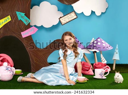 Little beautiful girl with long blond wavy hair in a blue silk dress in the scenery of Alice in Wonderland with white fluffy bunny cartoon fairy tale adventure