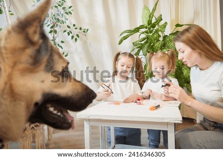 Young mother or babysitter, little daughter, sister teenager girl drawing at table and big dog shepherd looking. Family enjoying leisure at home