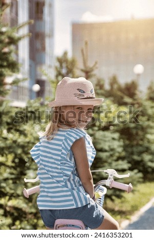 Rear view of little child in summer hat riding bike outdoors in city park and turned looking at camera. From behind cute adorable little blond girl enjoy riding small bicycle by path in playground