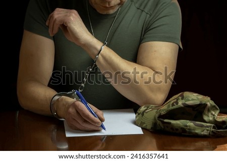 A military man sits at a desk, handcuffed, signing a document on white paper in a dimly lit room.