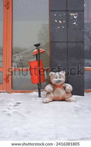 Teddy bear sitting in the snow near the building. Abandoned toy bear alone on the street in winter. A snow-covered freezing teddy bear. Royalty-Free Stock Photo #2416381805