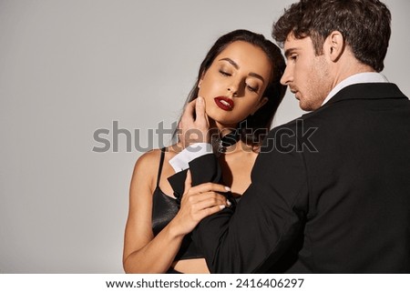 brunette young woman in black slip dress seducing handsome man in evening wear on grey background Royalty-Free Stock Photo #2416406297
