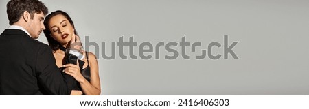 brunette young woman in black dress seducing handsome man in evening wear on grey background, banner Royalty-Free Stock Photo #2416406303