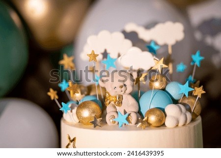 Blue and white cake decorated with a small bear, stars and clouds on sticks and gold and blue balloons on a background of inflatable white and green balloons