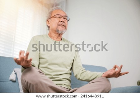 Lifestyle senior man sitting on sofa in living room holding hands in mudra practicing home yoga in lotus pose, Elderly relaxed man do physical exercises following healthy on stress free weekend