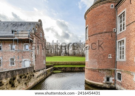 Moat surrounding 16th century Alden Biesen castle, between two buildings, one under renovation, green grass and bare trees in background, brick walls, cloudy day in Bilzen, Limburg, Belgium Royalty-Free Stock Photo #2416454431