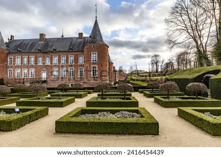 French garden with bushes and small trees, 16th century Alden Biesen Castle in background, bare trees, windows, brick walls, gable roof and circular towers, cloudy day in Bilzen, Limburg, Belgium Royalty-Free Stock Photo #2416454439