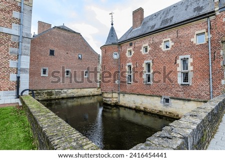 Part of moat surrounding 16th century Alden Biesen castle, seen from bridge, brick walls, circular towers and gable roof, building in background, cloudy day in Bilzen, Limburg, Belgium Royalty-Free Stock Photo #2416454441