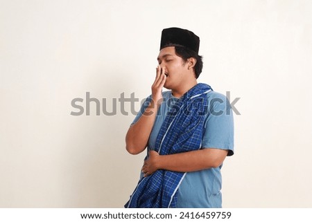 Sleepy young asian muslim boy with cap and scabbard standing while yawning Royalty-Free Stock Photo #2416459759