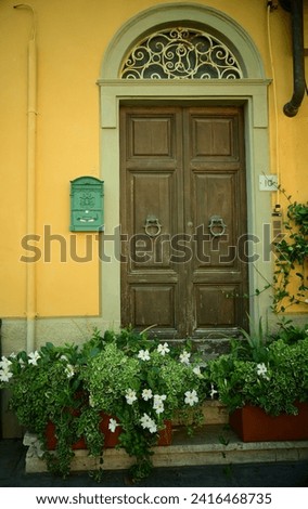 A portrait of an Italian, Tuscan, ornate, arched brown wooden gate on a mustard yellow building, with white flowers, a mailbox, and a portrait image. Cozy summer picture.

