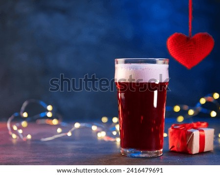 A glass of red beer, a small gift box and a red knitted heart hanging. Blue background, copy space