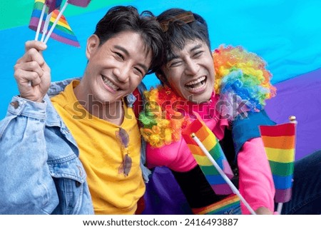 Two gay men sitting on rainbow flags on the ground, celebrate by waving flags to welcome the pride parades during Pride Month celebrations, LGBTQ concept