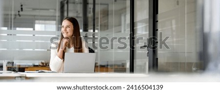 A contemplative businesswoman enjoys a moment of reflection while working on a laptop, her gaze suggesting strategic thought and planning in a spacious office setting. The panoramic view, banner Royalty-Free Stock Photo #2416504139