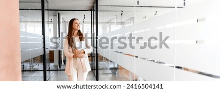 A cheerful businesswoman in an ivory suit walks through a modern glass office corridor, carrying a laptop, her smile suggesting a successful meeting or a job well done, panoramic view, banner Royalty-Free Stock Photo #2416504141