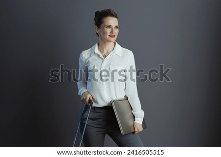 pensive modern middle aged business woman in white blouse with laptop and trolley bag against gray background.