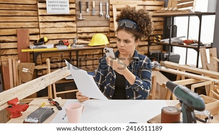 Young woman examines documents in a carpentry workshop, surrounded by tools and wood. Royalty-Free Stock Photo #2416511189