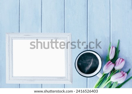 Blank empty picture frame with hot coffee and tulip flowers over blue rustic background with spring flowers. Table top view.