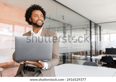 Poised African American businessman in a taupe suit holds his laptop and looks off into the distance, reflecting confidence and vision in a modern office setting standing in bright and open workspace Royalty-Free Stock Photo #2416529377
