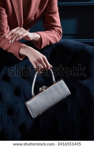 Silver metallic sparkling purse held by woman hands on a blue velour armchair background. Creative shiny woman hand bag studio photography. Royalty-Free Stock Photo #2416551435