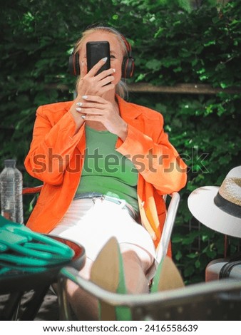 A  stylish blonde woman in a bright orange jacket with a green bag, headphones holds a smartphone in front of her face, having fun communicating, sits with her legs stretched out on a chair 
