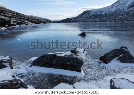 A winter landscape of rocks locked in the ice of Dubh Loch in the Scottish Highlands. The smooth ice stretches into the distance with steep, snow dusted mountains rising on either side.