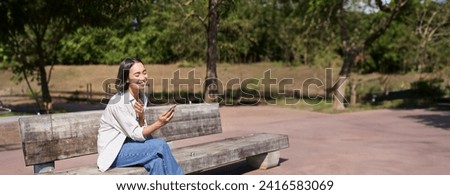 Stylish young asian girl, student sitting in park with smartphone, using telephone, waiting for someone while sitting on bench. Royalty-Free Stock Photo #2416583069