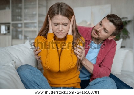 Disappointed woman sitting on sofa, closing ears with hands, doesnt want to hear man justifying, expaining disagreement in relations. Tension, emotional confrontation, misunderstanding in family life Royalty-Free Stock Photo #2416583781