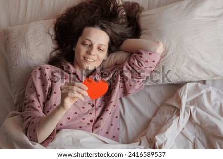 happy woman reading congratulations on postcard in morning. red card in form of heart for Valentine's Day. female in pajamas lies and enjoys her vacation. gift from lover, tradition of giving gifts