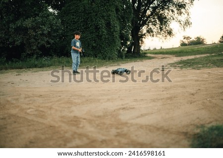 Picture of a boy operating an electric race car toy on a dirt road in the countryside. He is holding a large remote controller while there are various trees behind him.