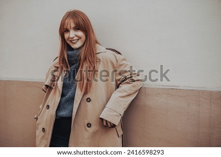 Portrait of a fashionable young woman in trendy outfit leaning on a grungy wall and smiling at the camera. A stylish lady is posing on a city street downtown and smiling at the camera. Copy space.