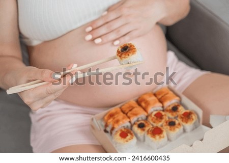 A pregnant woman sits on the sofa and eats rolls from a box. Food delivery. Close up of belly.  Royalty-Free Stock Photo #2416600435