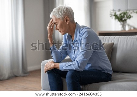 Depressed european senior lady suffering from headache touching head, sitting on sofa in modern living room at home, thinking about problems, having mental health issues and depression Royalty-Free Stock Photo #2416604335